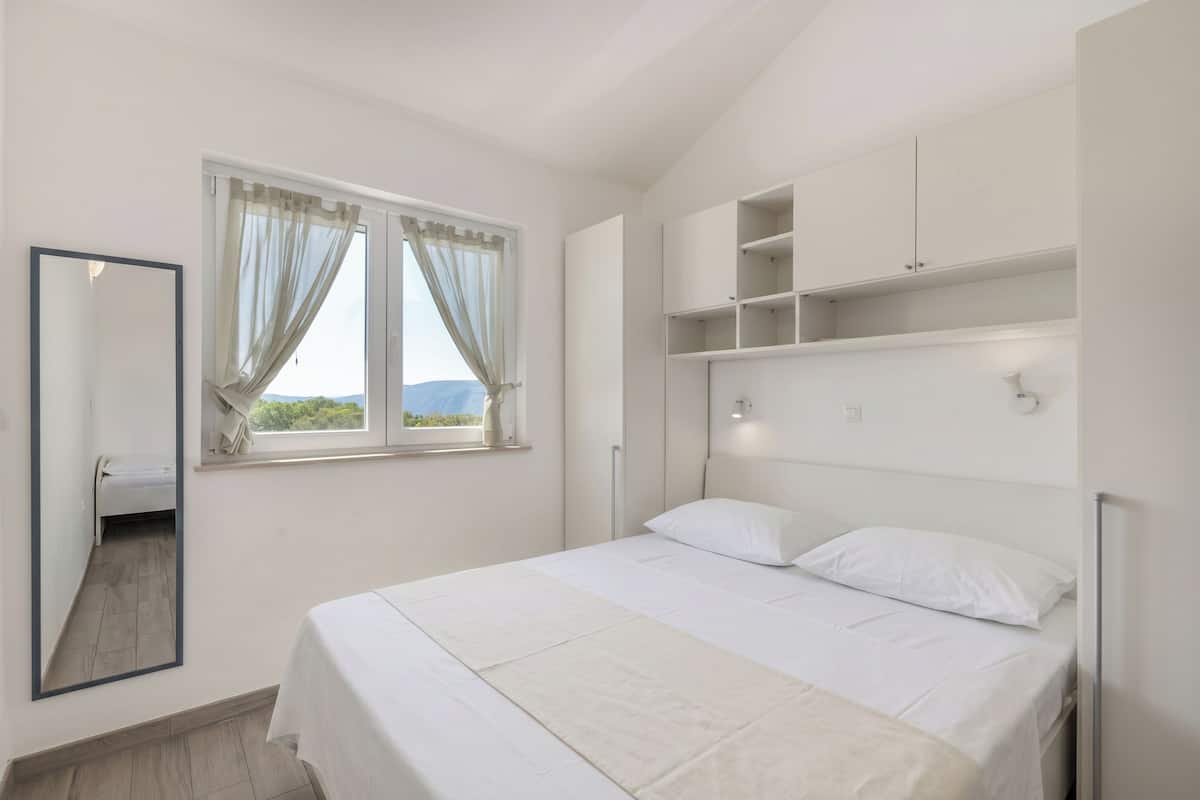A bedroom with a bed, a window with curtains, a mirror, and built-in shelving.