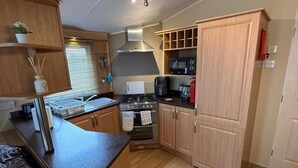 A compact kitchen with a stove, microwave, and sink, wooden cabinets, and a window with blinds. - Cabin Caravan Sleeps 6 With Parking (Abergele)