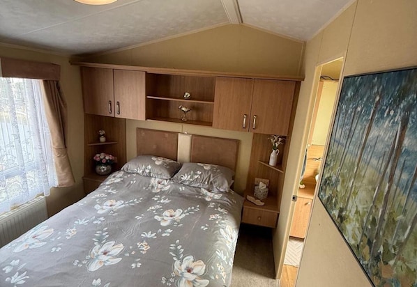 A bedroom with a bed, wooden cabinets, a painting, and a window with curtains. - Cabin Caravan Sleeps 6 With Parking (Abergele)
