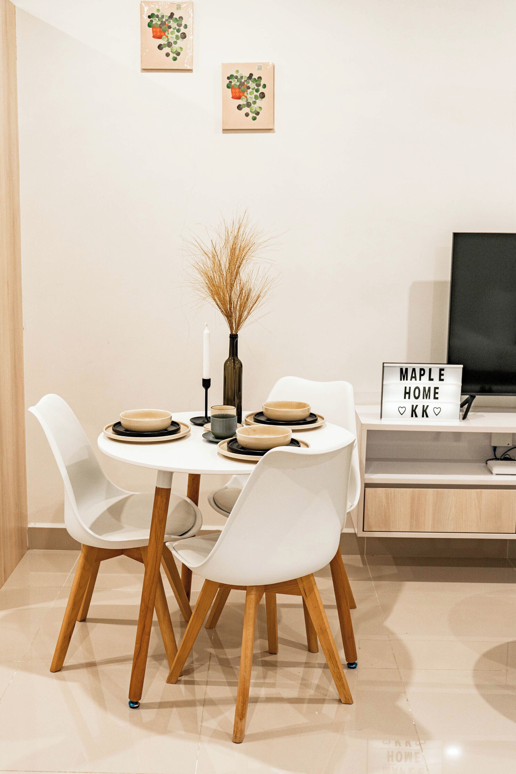 Un comedor con una mesa redonda, sillas blancas, un jarrón con plantas secas y un letrero en la pared.