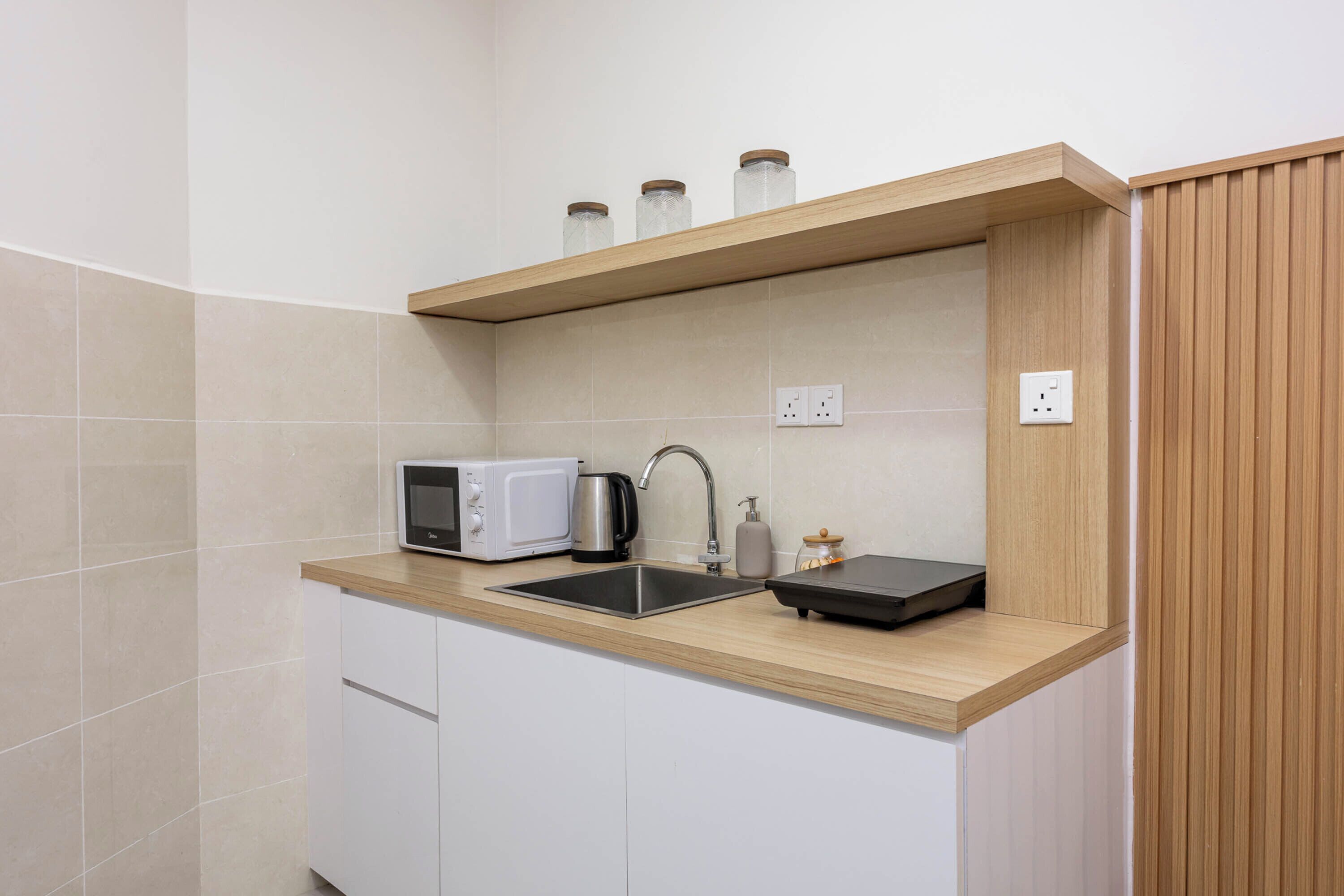 A compact kitchen with a microwave, kettle, sink, and wooden shelf with jars.