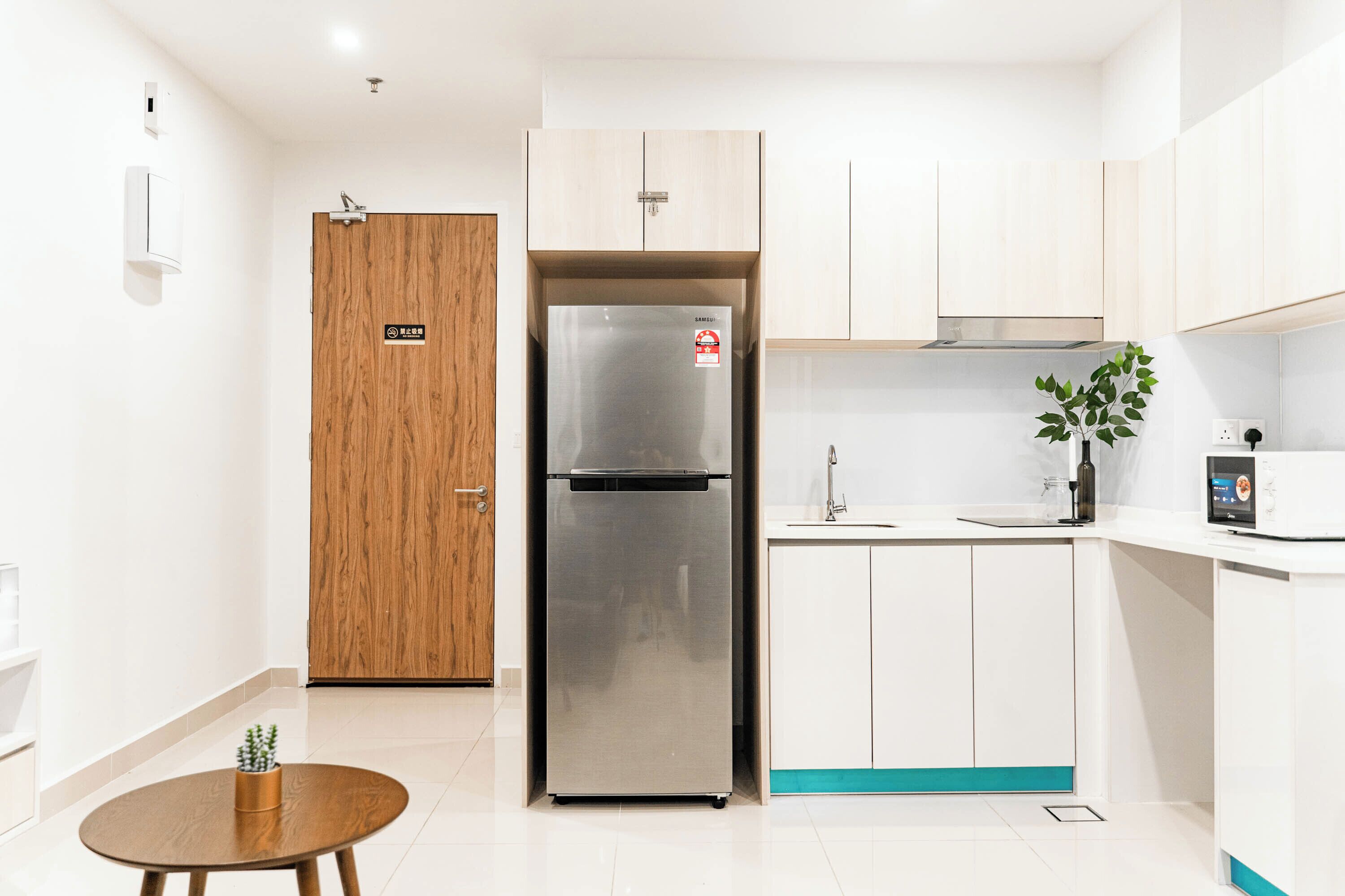 A modern kitchen with a stainless steel refrigerator, white cabinets, a wooden door, and a small round table with a plant.