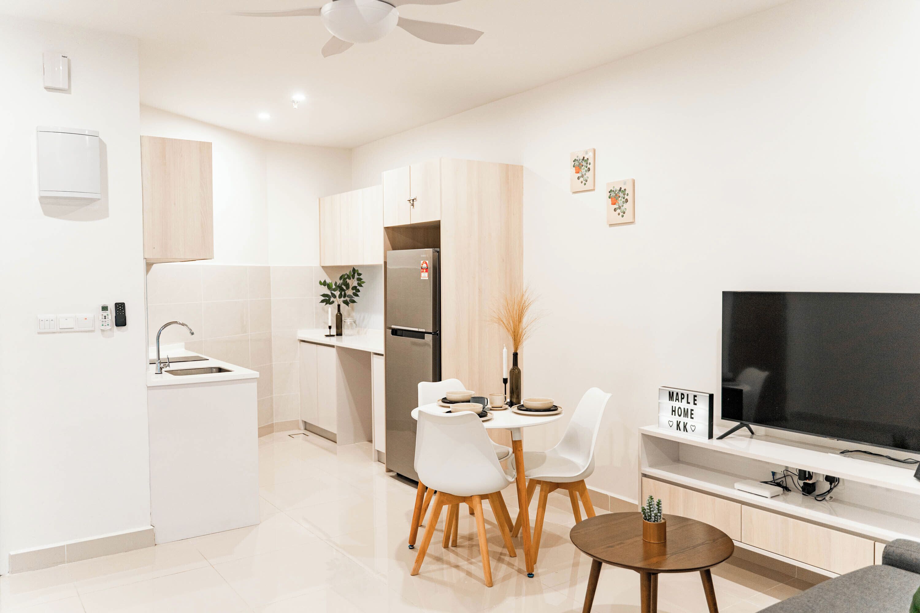 Una cocina moderna con armarios blancos, horno empotrado y refrigerador. Hay una zona de comedor con sillas blancas y una mesita pequeña. Un televisor de pantalla plana está montado en la pared sobre una consola blanca.