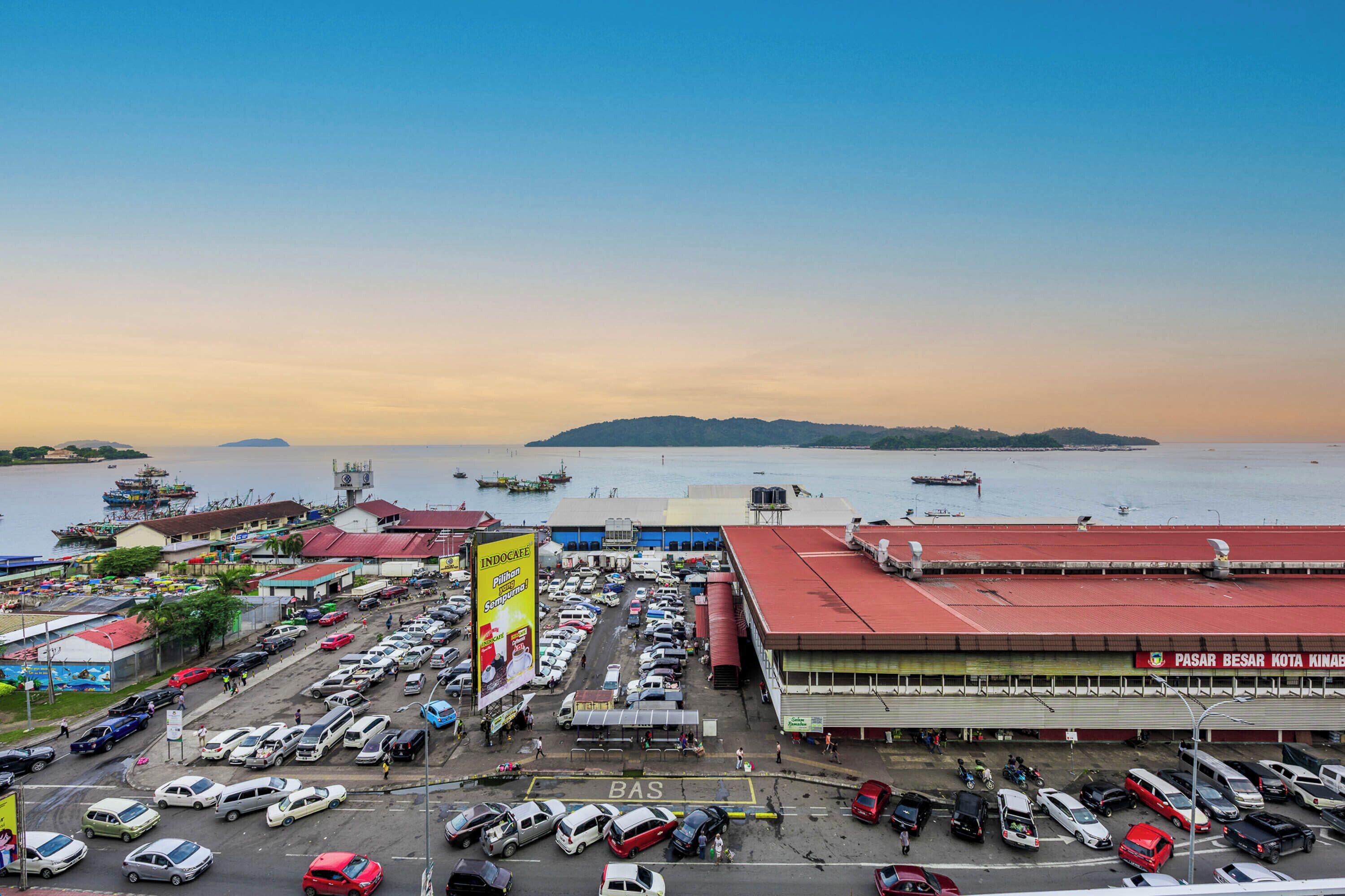 A parking lot with multiple cars, a large building with a red roof, and a body of water with boats in the background.