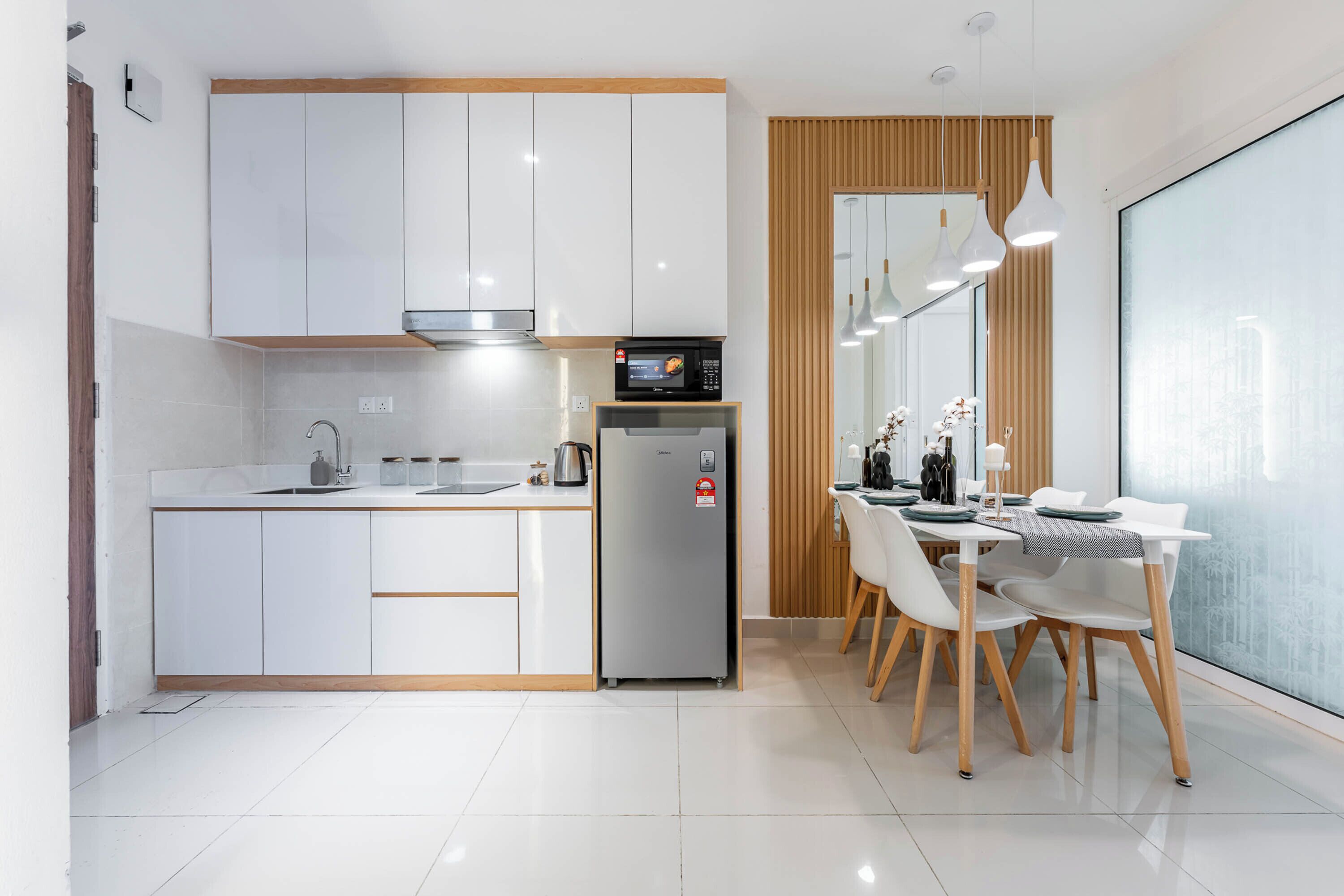 A modern kitchen with white cabinets, a built-in microwave, and a refrigerator. There is a dining area with white chairs and a table.