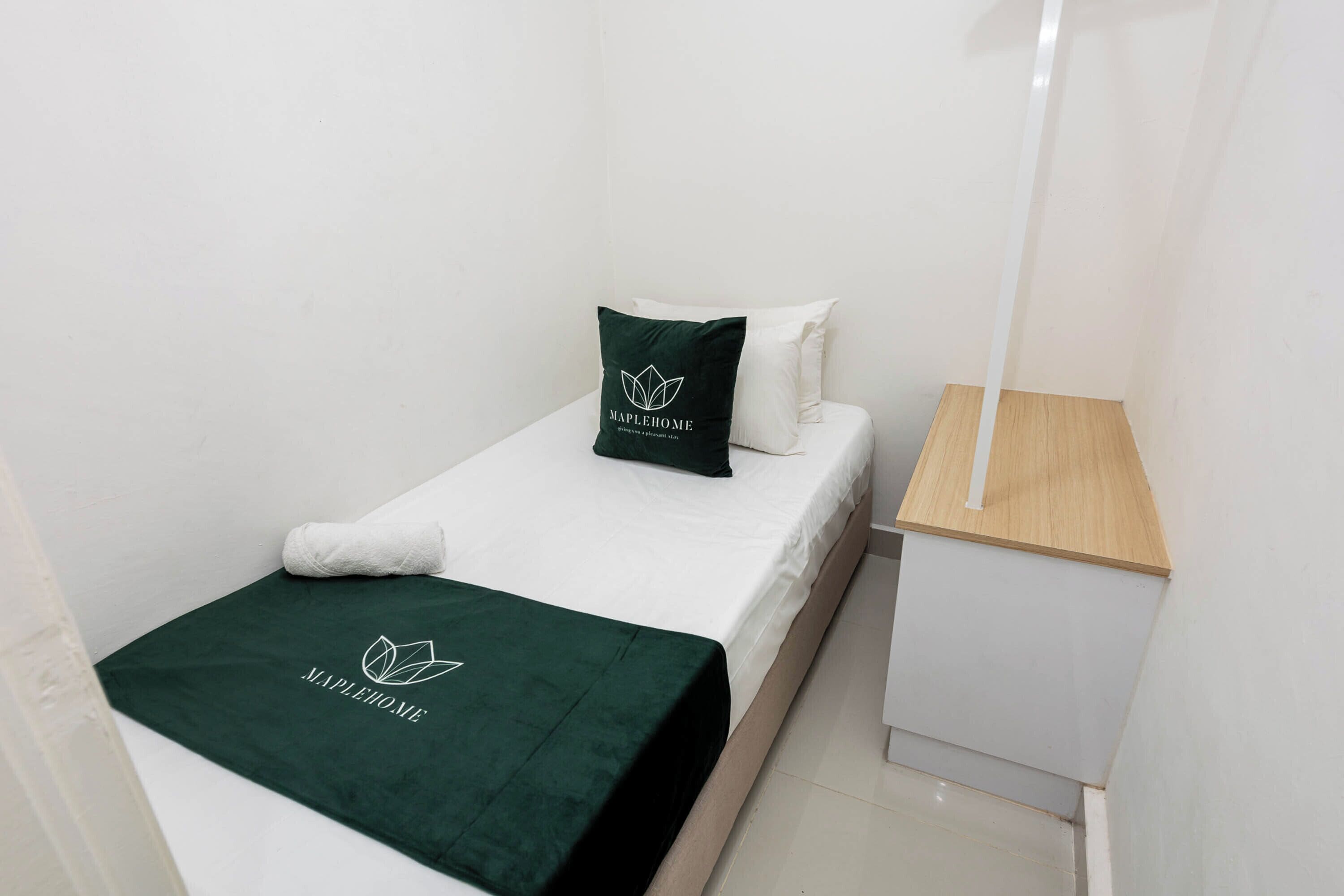 Una cama individual con colcha verde y almohada, una mesita de noche blanca y una pequeña mesita auxiliar de madera.