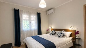 Een slaapkamer met een bed, nachtkastjes, een bankje, een raam met gordijnen, een airconditioning en een wandlamp.