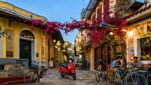 Una calle adoquinada con bicicletas, un scooter rojo y personas sentadas en mesas al aire libre.
