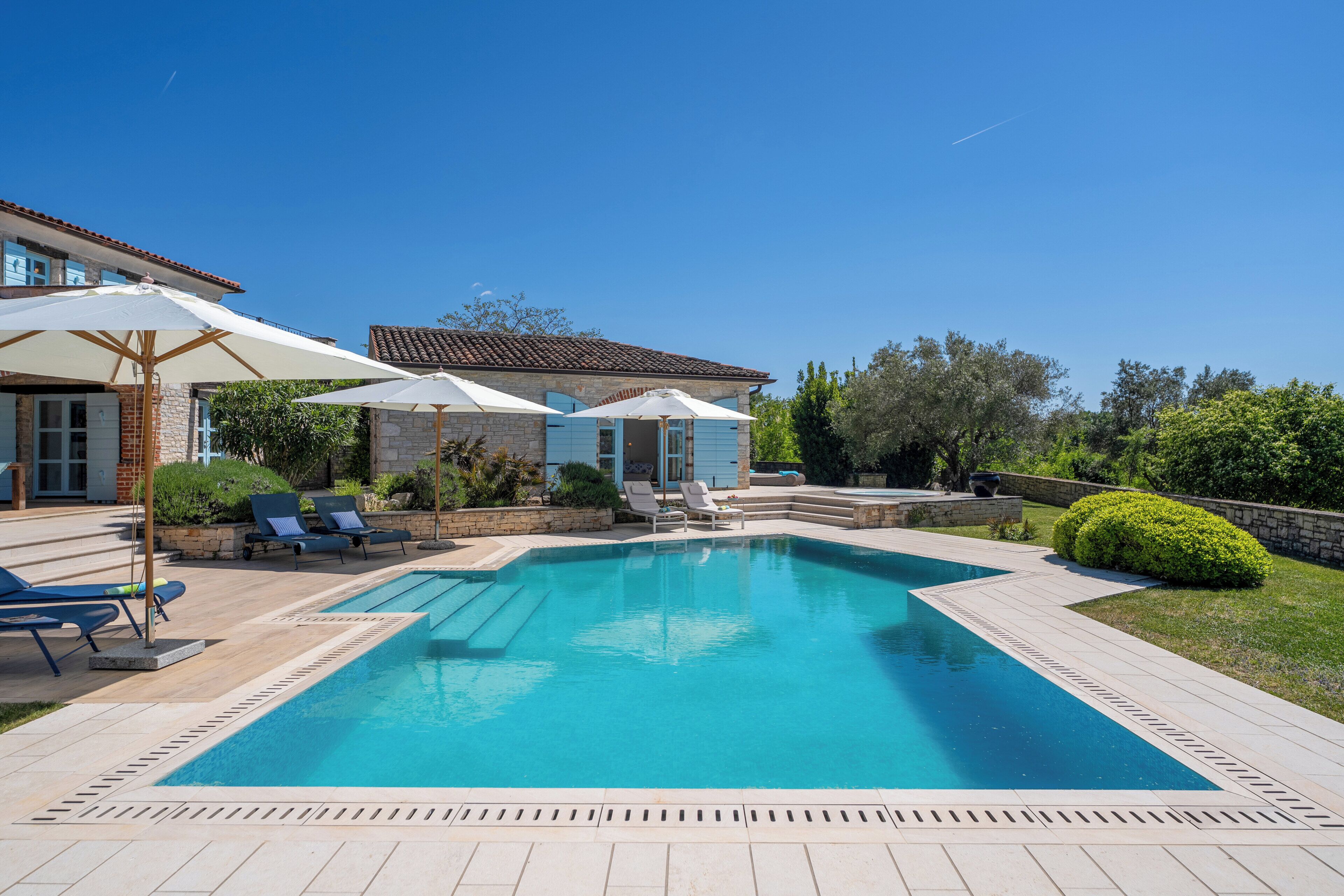 A swimming pool with lounge chairs and umbrellas, surrounded by a landscaped garden and a house with a tiled roof.