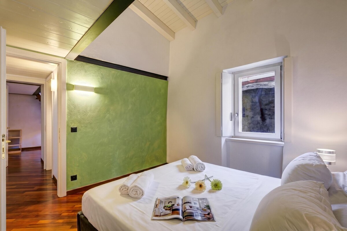Camera d'albergo con un letto, una finestra vista, una parete verde decorativa e una rivista sul letto.