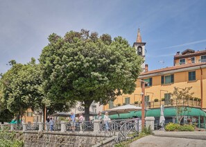 Une place bordée d’arbres, avec un bâtiment doté d’un clocher, des places assises en plein air et un mur en pierre.