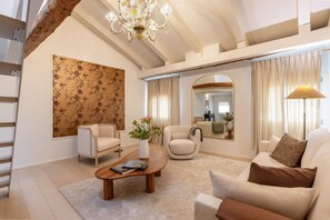 Ein geräumiges Wohnzimmer mit hoher Decke, einem Chandelier, einer großen Blumen-Tapete, einem runden Spiegel und einem Sofa mit braunem Zierkissen.