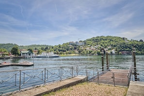 A waterfront dock with a boat, a pier with a metal railing, and buildings on a hillside in the background. - Holiday Home 'Unit D' by the Lake with Large Garden, Wi-Fi and Air Conditioning (Laveno-Mombello)