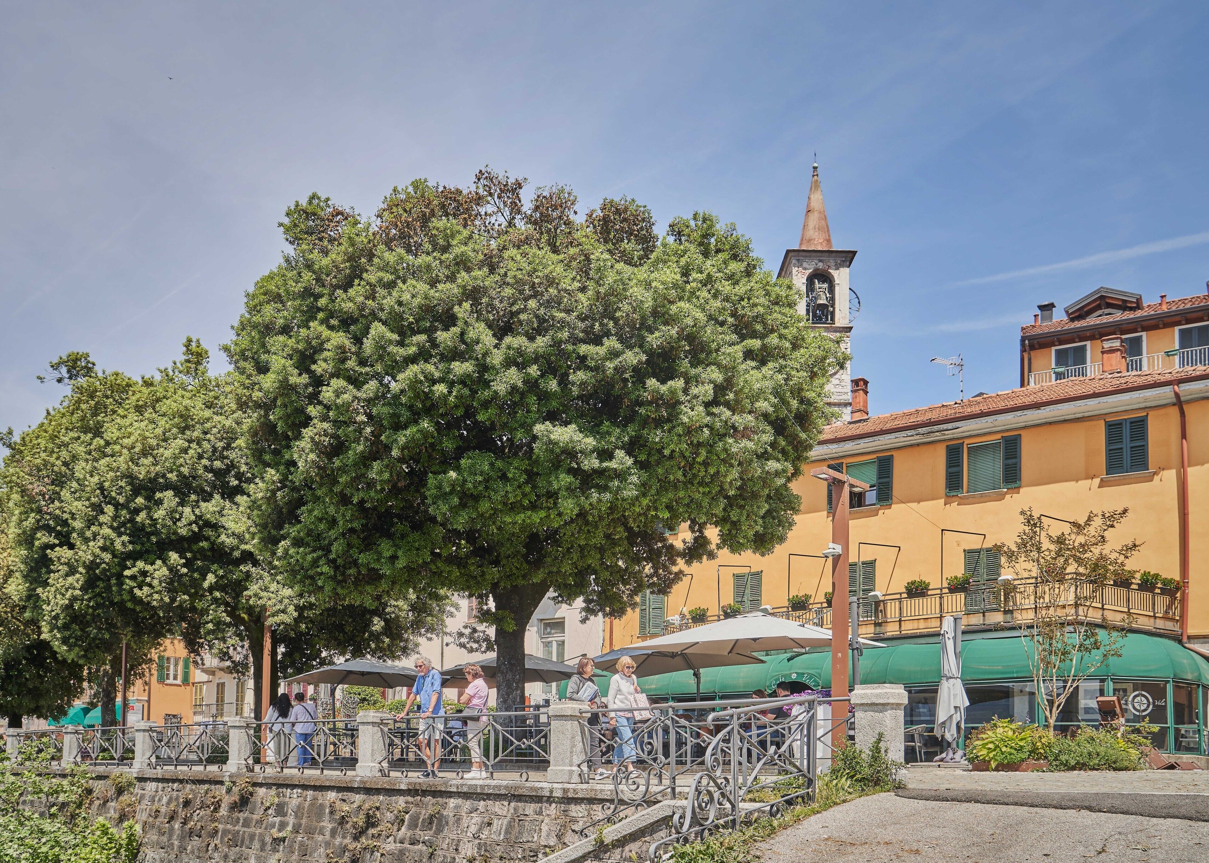 Una plaza arbolada con un edificio que cuenta con una torre de campanas, mesas al aire libre y un muro de piedra.