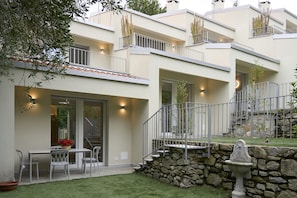 Um edifício moderno de vários andares com varandas, uma parede de pedra e uma pequena área de jardim com mesa e cadeiras.