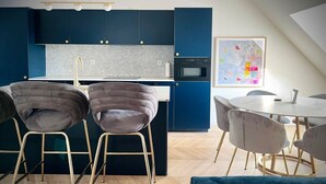 Et moderne køkken med mørkeblå skabe, en hvid bordplade og en indbygget ovn. Der er to grå polstrede stole med gyldne ben, et rundt spisebord med hvide stole og en blå sofa.