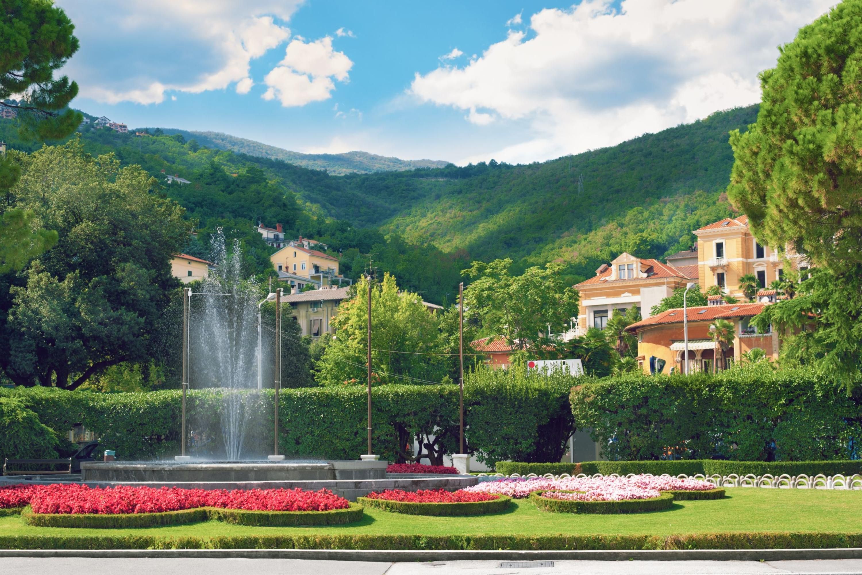 Ein Springbrunnen, umgeben von gepflegten Blumenbeeten, mit Gebäuden im Hintergrund.
