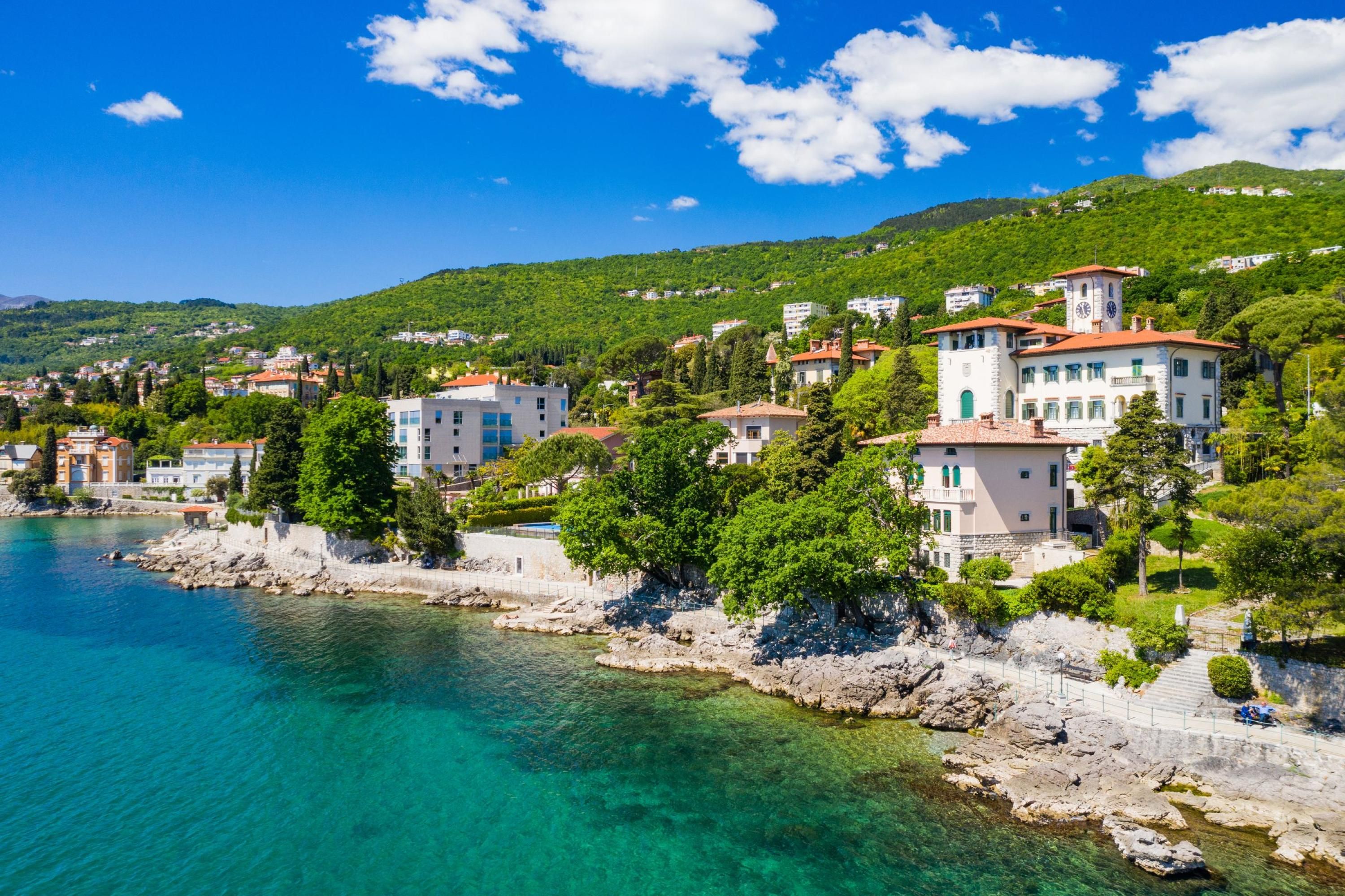 A coastal town with buildings along the waterfront, clear blue waters, and green hills in the background.
