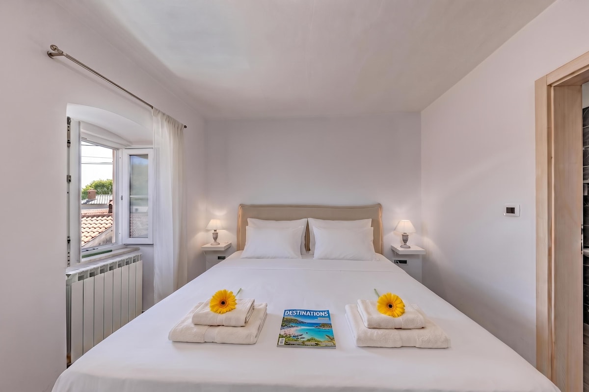 Ein Hotelzimmer mit einem Bett, zwei Nachttischlampen, einem fensterbelaubten Fenster, einer Heizung und einer Zeitschrift auf dem Bett.