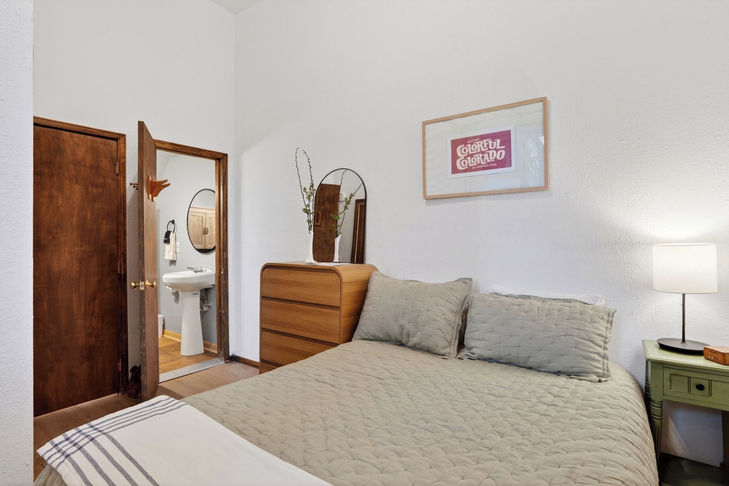 Un dormitorio con una cama, un aparador, un espejo, un cartel enmarcado y una mesita de noche con una lámpara.
