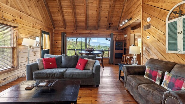 A wooden cabin interior with a sofa, a table, and a view through a large window. - Covered Deck + Fire Pit + Mountain Views | Near Smokies | Isaiah's Cabin (Townsend)