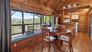 A wooden cabin with a dining table and chairs, a kitchen area, and large windows offering a view of the outdoors. - Covered Deck + Fire Pit + Mountain Views | Near Smokies | Isaiah's Cabin (Townsend)