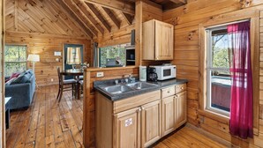 A wooden cabin interior with a kitchen, dining area, and a window with curtains. - Covered Deck + Fire Pit + Mountain Views | Near Smokies | Isaiah's Cabin (Townsend)