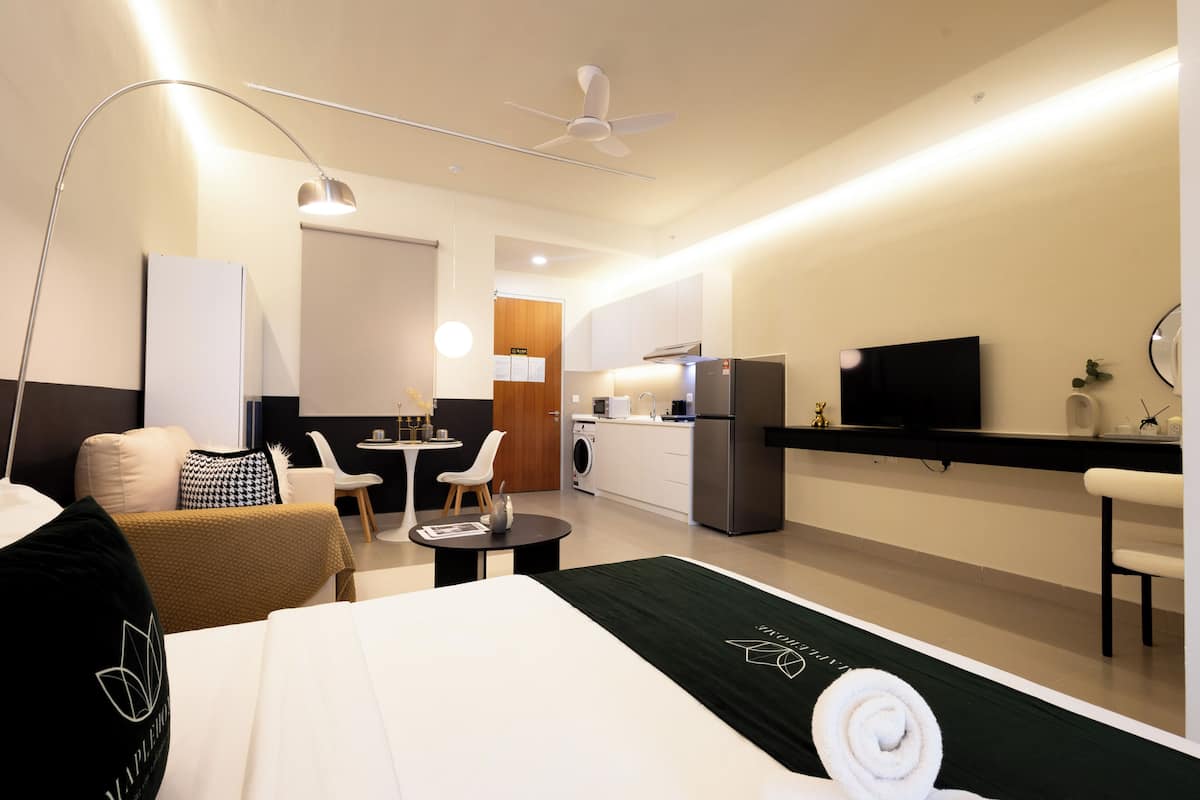 Habitación de hotel con cama, escritorio, silla, mesita, televisor y zona de cocina con refrigerador, además de ventilador de techo.