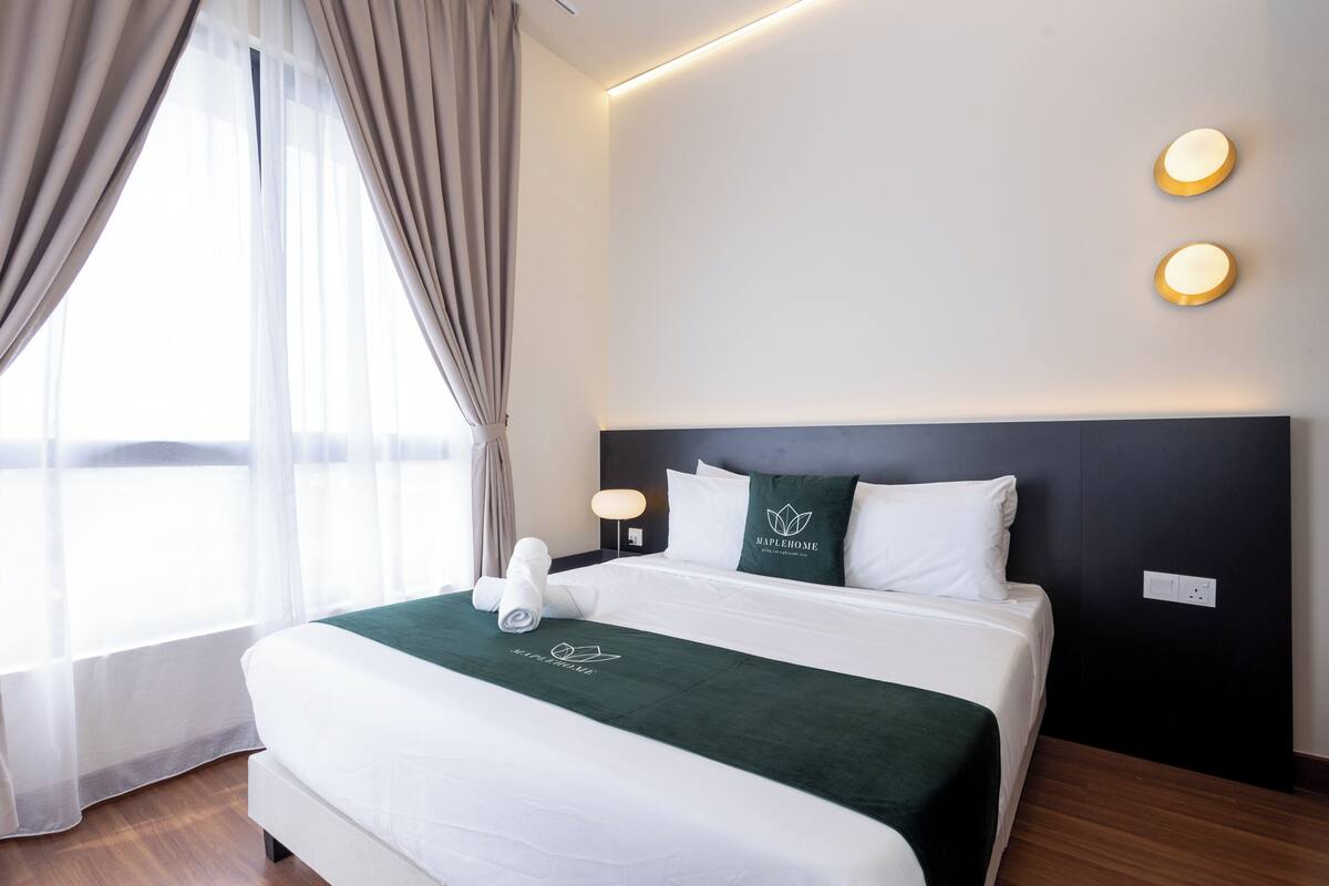 Una cama bien hecha con cabecera verde, ropa de cama blanca y una toalla. Hay dos luces fijadas a la pared y una ventana con cortinas de gasa.