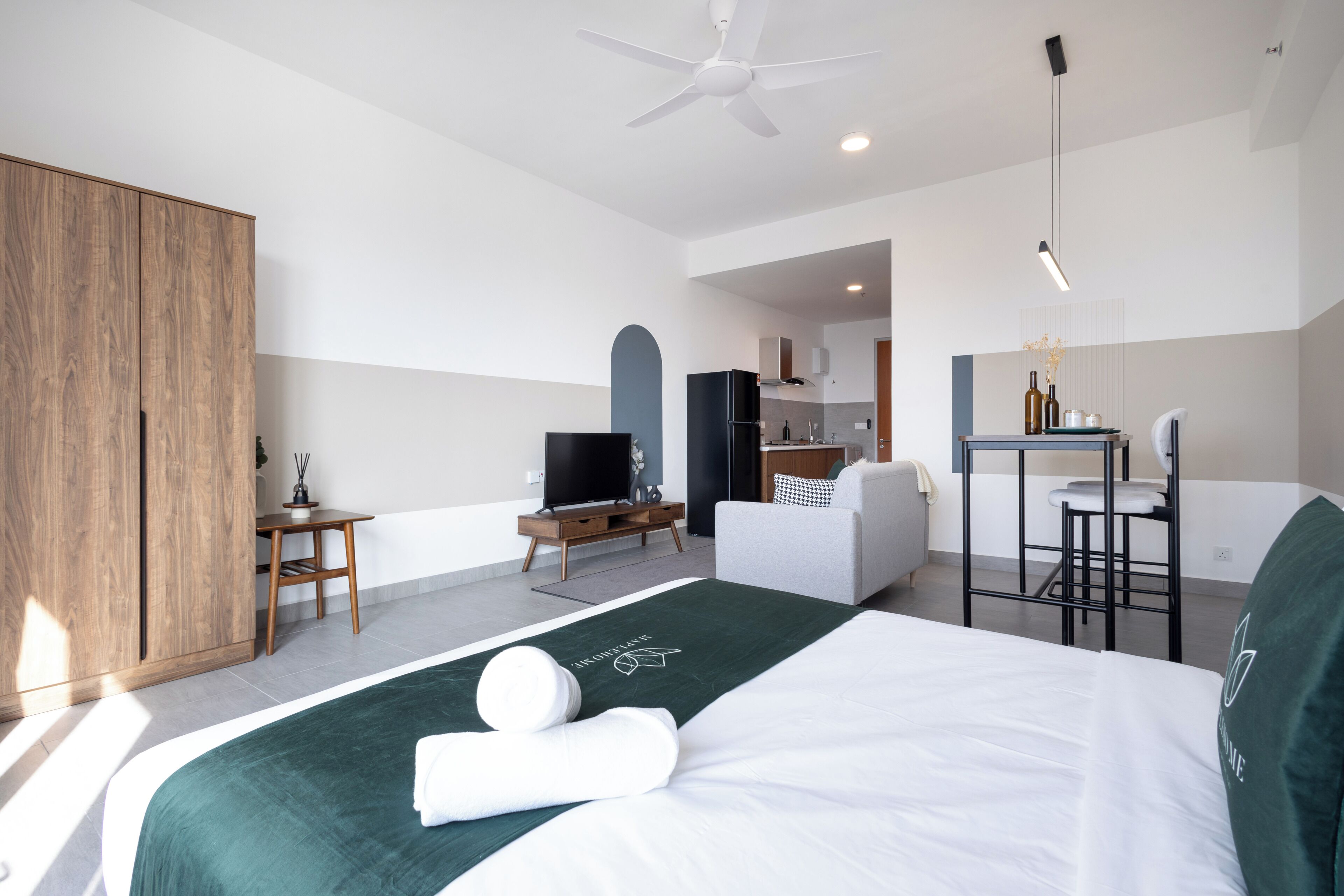 A modern hotel room with a bed, a desk, a television, a wardrobe, and a kitchenette.
