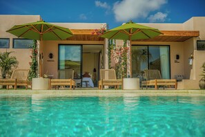 Zona de piscina con sillones de descanso y sombrillas, una piscina cristalina y la entrada de un edificio con puertas de vidrio.