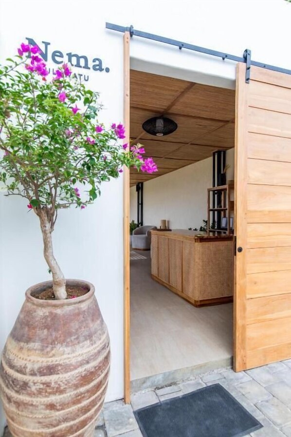 Une porte en bois donne sur une pièce où se trouvent un banc en osier et une plante en pot.