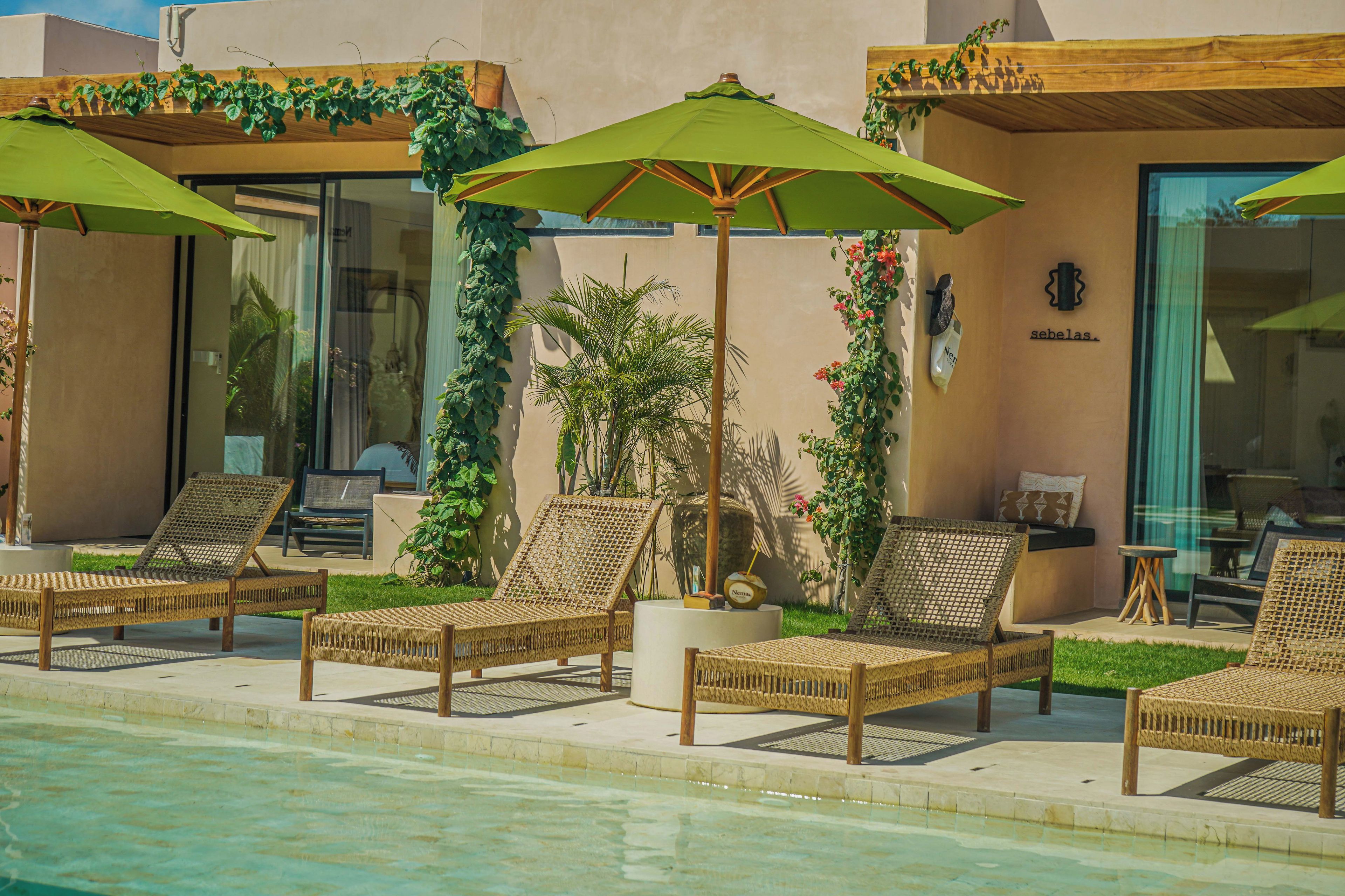 Un espace aménagé au bord de la piscine, avec des fauteuils de relaxation en osier et des parasols verts, un palmier, et un mur orné d’une enseigne « sebelas ».