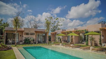 Una piscina con sillas de descanso y sombrillas, rodeada de un jardín ajardinado y un edificio con grandes ventanales.