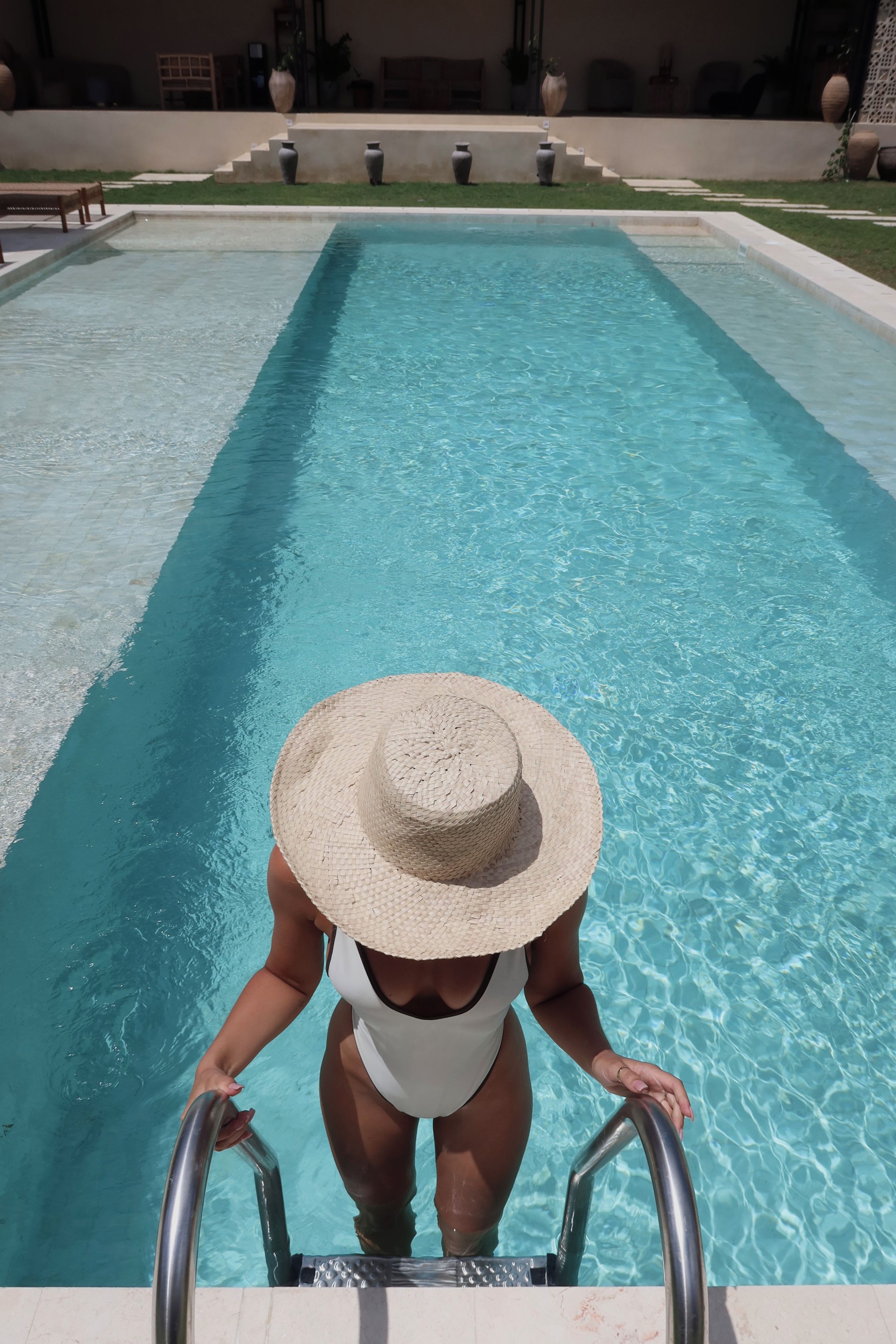 Ein Swimmingpool mit klarem, blauem Wasser, eine Person im Badeanzug und mit Strohhut, sowie ein Poolbereich mit Stufen zum Wasser.