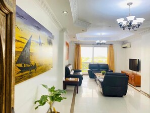 A living room with a large painting, a chandelier, a television, and a view of the outdoors. - Oysterbay Pool View Two Bedroom (Dar es Salaam)