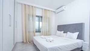 A modern bedroom with a large bed, white linens, a grey headboard, and a window with sheer curtains. - sandra 11 (La Marsa)