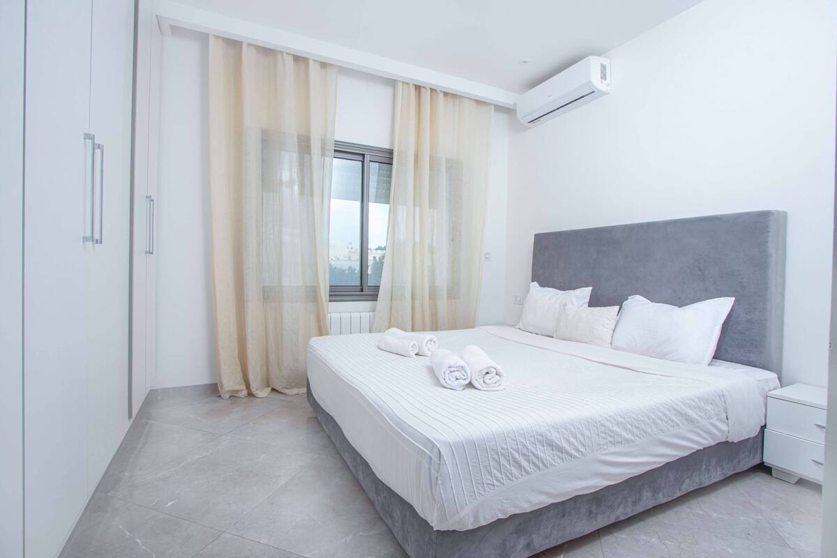 Quarto moderno com uma cama grande, roupa de cama branca, cabeceira cinza e uma janela com cortinas leves.