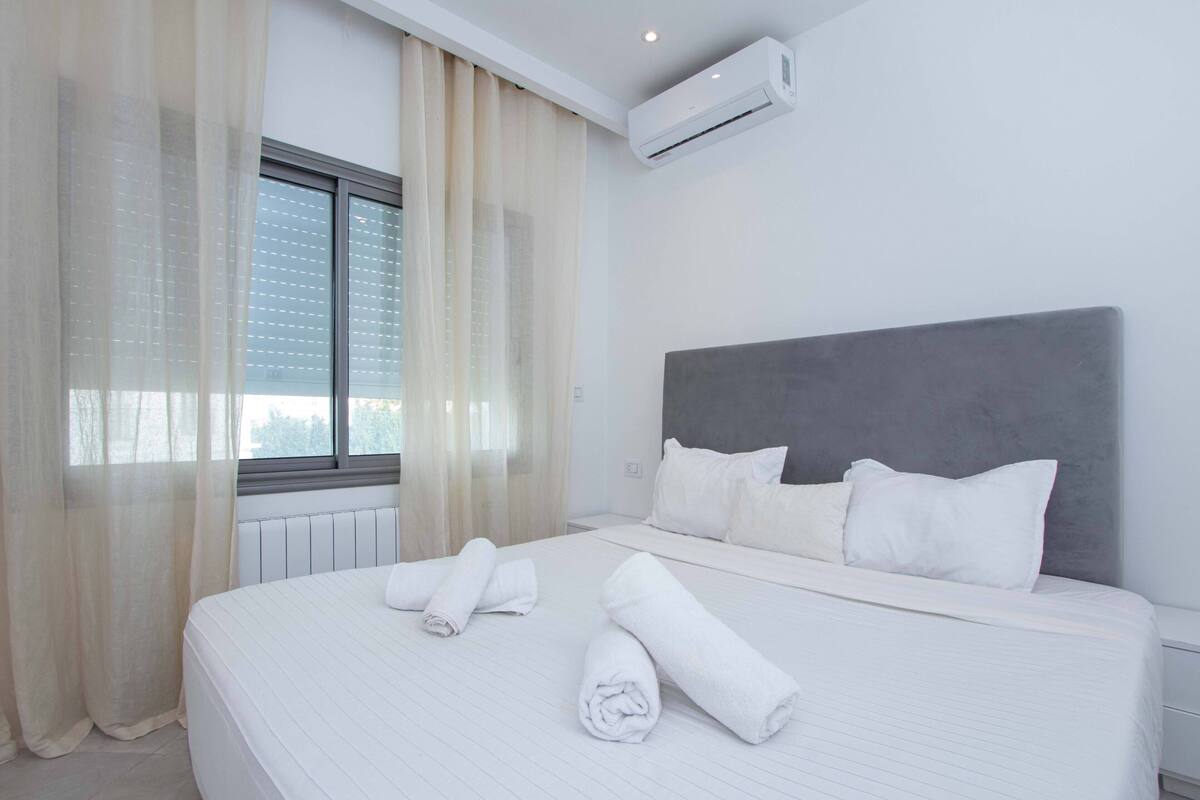 Quarto de hotel com uma cama grande, roupa de cama branca, cabeceira cinzenta e uma janela com cortinas translúcidas.