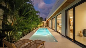 A swimming pool with lounge chairs, surrounded by tropical plants, and a modern building with large windows. - Villa Malaikat (Canggu)