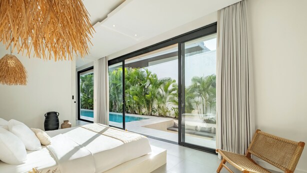 A modern bedroom with a large bed, a wicker chair, and a view of a swimming pool. - Villa Malaikat (Canggu)