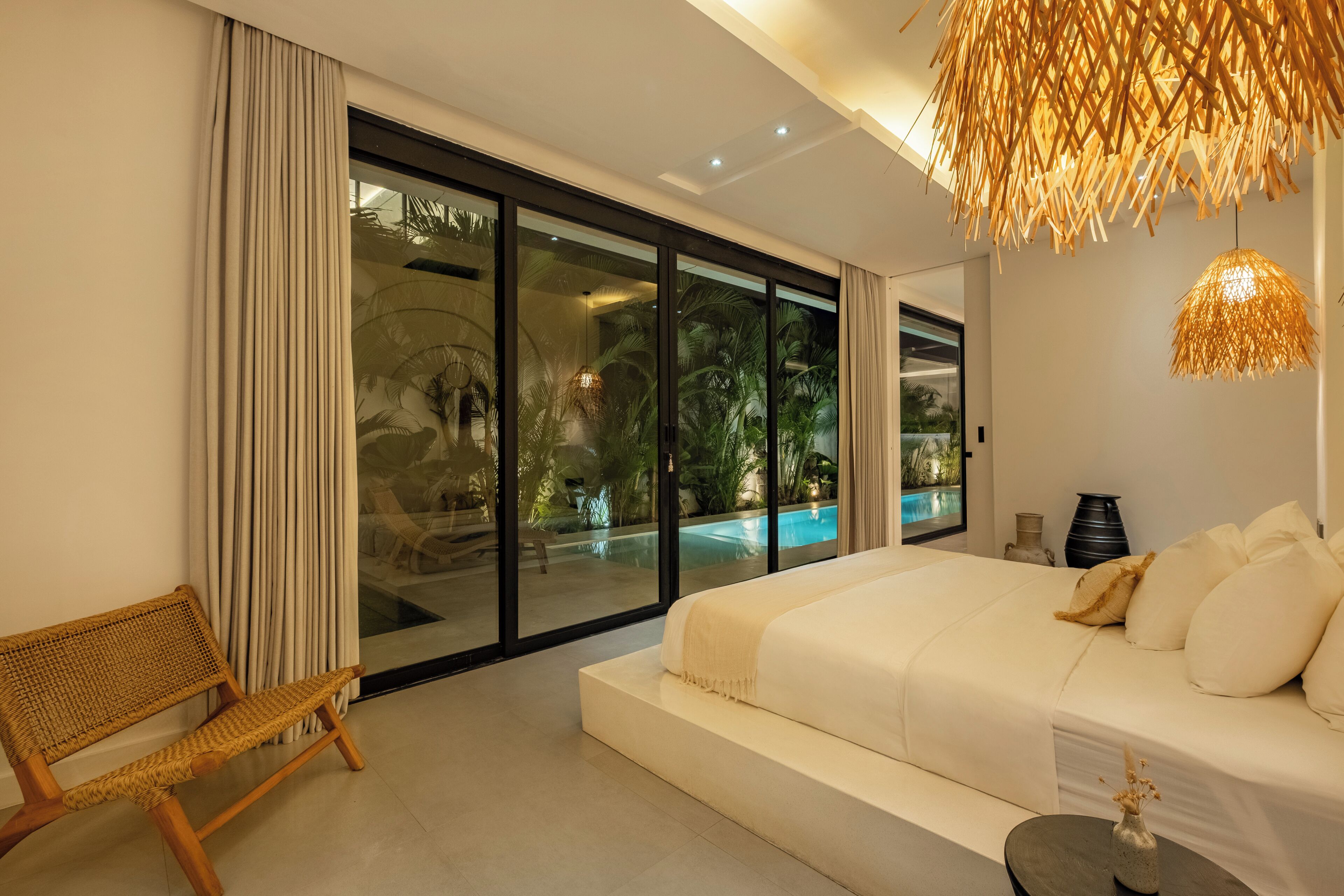 Un dormitorio moderno con una cama grande, un sofá, una silla de mimbre, una mesita y vista a una piscina a través de puertas de vidrio.