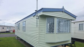 A mobile home with a flat roof, two windows with lace curtains, and a small porch area.