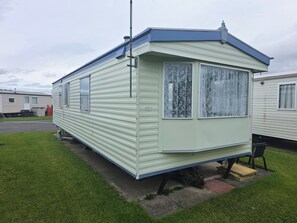 Une maison mobile dotée d’un toit plat, de deux fenêtres ornées de rideaux en dentelle et d’un petit porche.