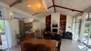 Un amplio salón con una gran mesa de comedor de madera, diversas opciones de asiento, chimenea y vistas a través de grandes ventanales.