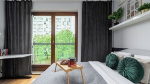 Une chambre à coucher avec un lit, une table de chevet avec un plateau, une fenêtre avec des rideaux et une vue sur des immeubles et des arbres.