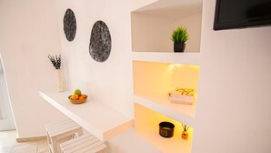 A kitchen with a white countertop, a bowl of fruit, a potted plant, and a shelf with a candle and a small container. - Windmills Suite by Opus (Mykonos)