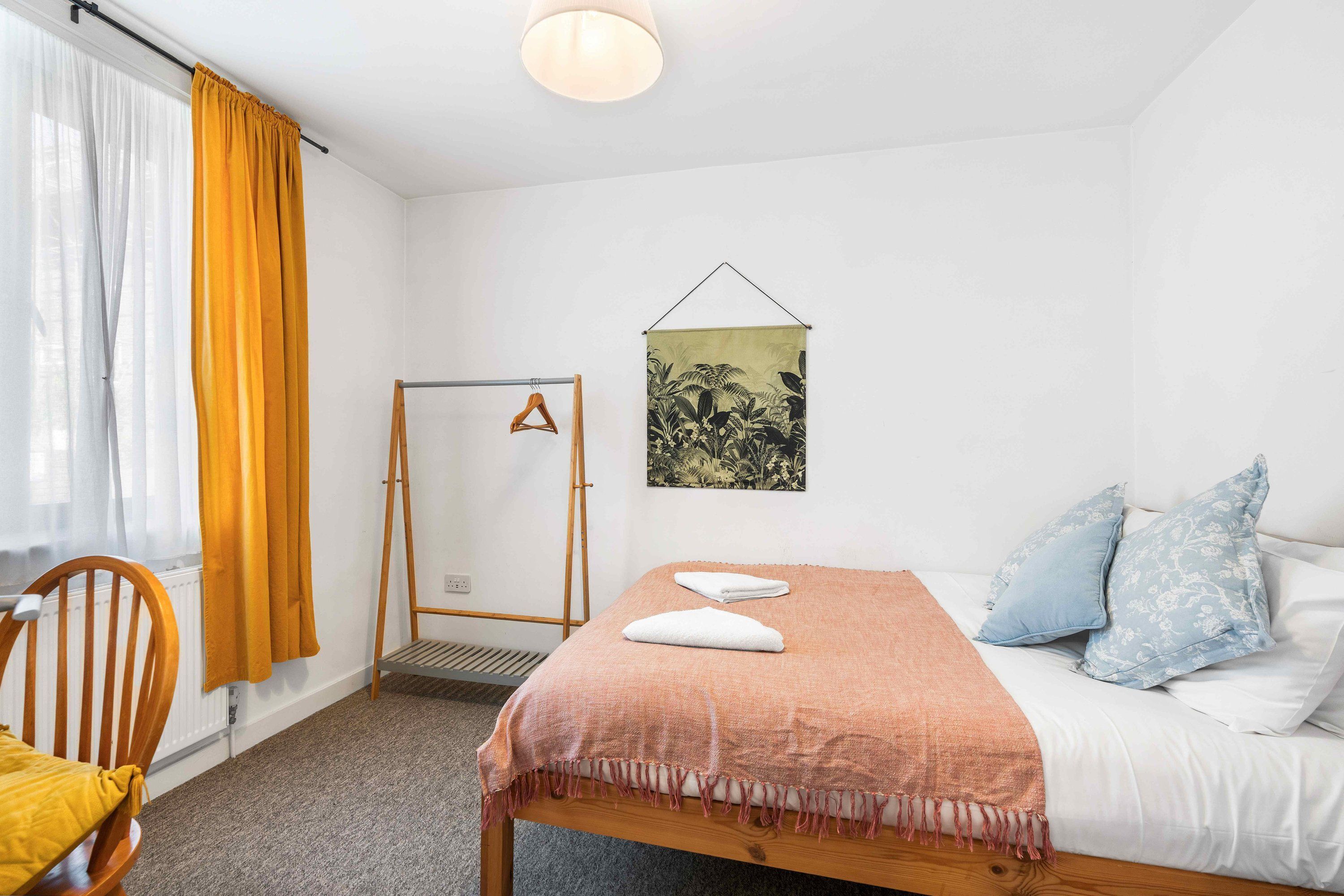 A bedroom with a bed, a chair, a radiator, a window with curtains, and a wall-mounted artwork.