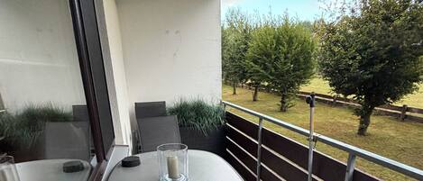 Un balcón con mesa y sillas, un vaso de agua y vistas a un área verde con árboles.