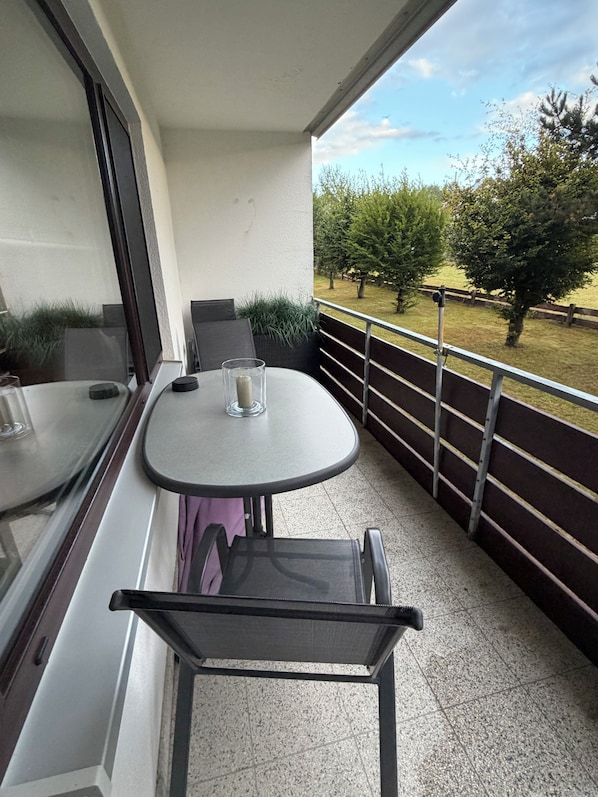 A balcony with a table and chairs, a glass of water, and a view of a grassy area with trees.